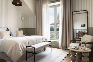 Un dormitorio con cama, banco, mesa redonda, silla y vista a un edificio a través de una ventana.