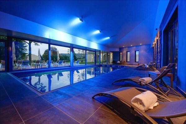 A modern indoor swimming pool with clear glass walls, lounge chairs, and a view of the outdoors. - Hotel Spa La Cueillette (Meursault)