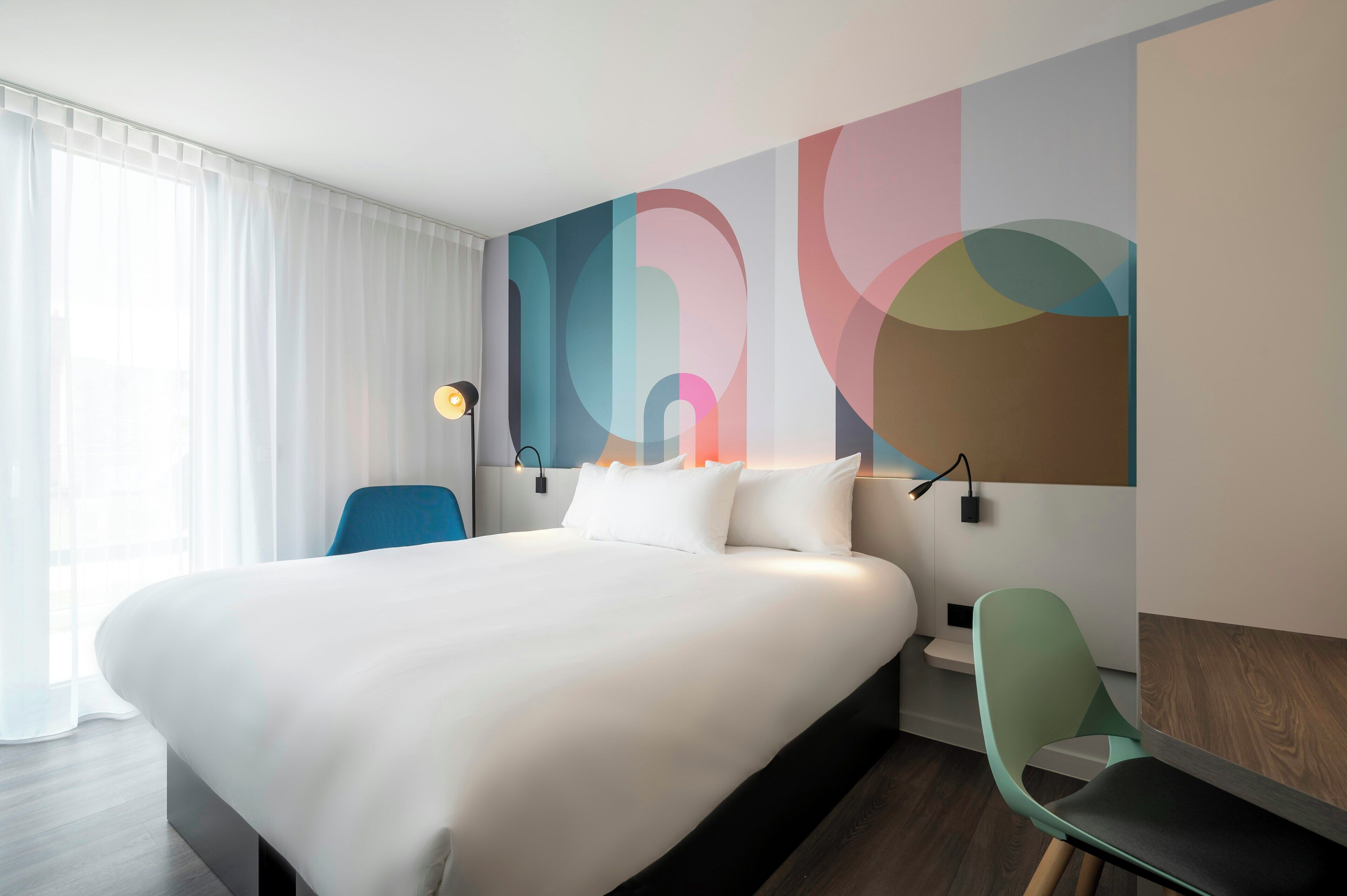 A hotel room with a large bed, a desk, a chair, and a wall with abstract art.
