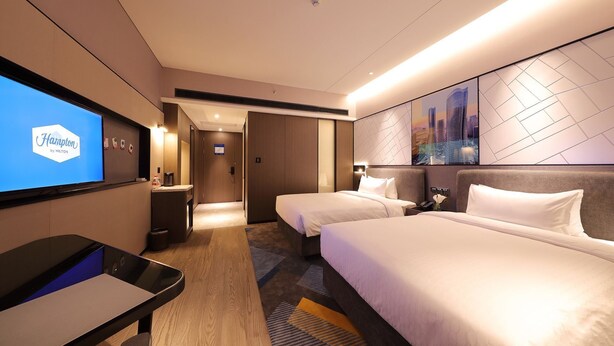 Habitació d'hotel amb dos llits, un escriptori, un televisor que mostra 'Hampton by Hilton' i una obra d'art amb un paisatge urbà a la paret.