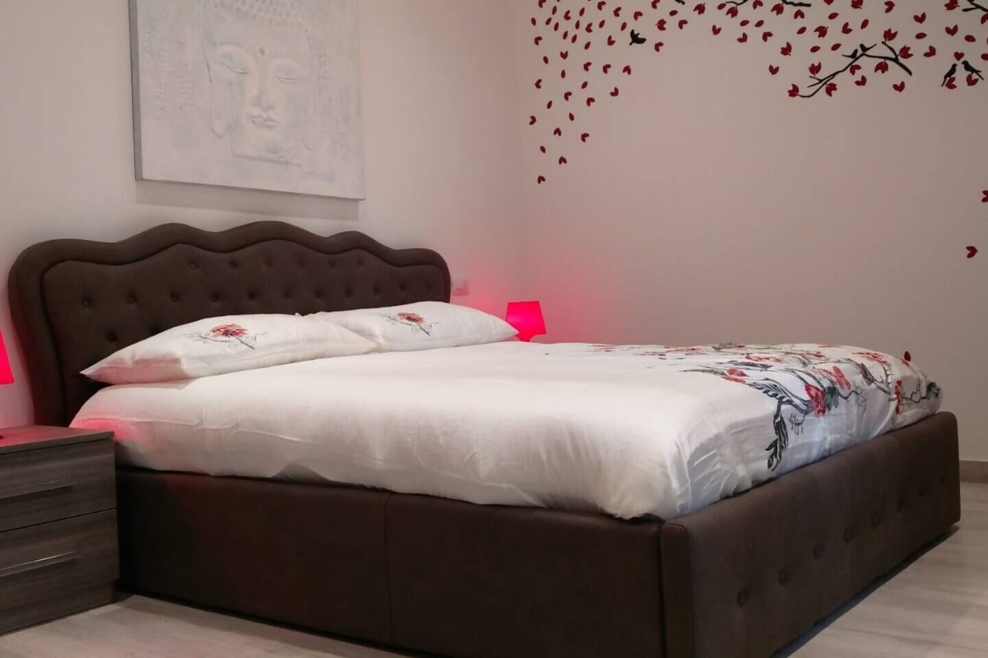 A neatly made bed with a floral bedspread and pillows, a brown upholstered headboard, a bedside table with a lamp, and a wall decoration of birds and leaves.