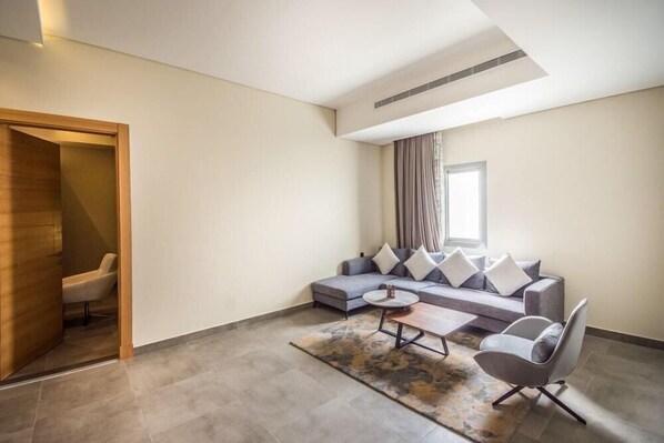 Una sala moderna con un sofá modular, una mesa de centro y una butaca. Se observa una puerta de madera que da a otra habitación, y una ventana con cortinas.