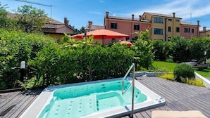 Una terraza de madera con un jacuzzi, rodeada de vegetación y un edificio con techo rojo.