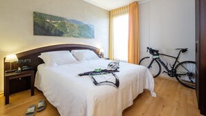 Habitación de hotel con una cama bien hecha, una bicicleta, pantuflas y un cuadro en la pared.