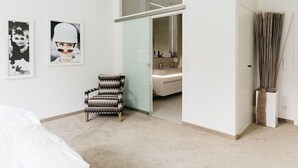 Une chambre d’hôtel moderne avec un lit, un fauteuil à motifs, une salle de bain avec un miroir et un lavabo, et deux tableaux encadrés accrochés au mur.