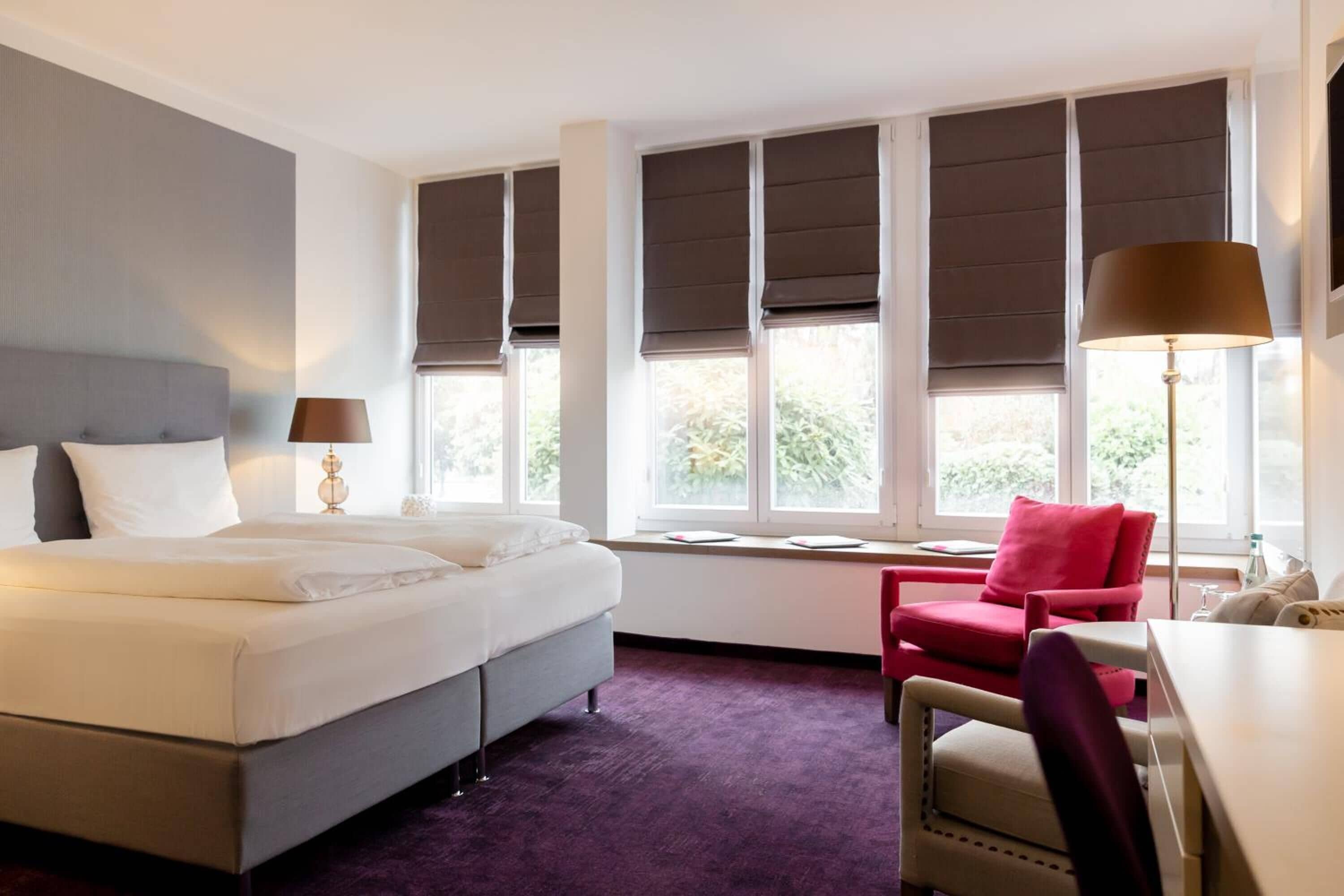 Ein Hotelzimmer mit Bett, Schreibtisch, Stuhl und großen Fenstern mit Jalousien.