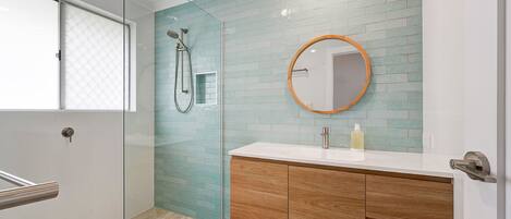 A bathroom with a glass shower enclosure, a wooden vanity, a round mirror, and a tiled backsplash.