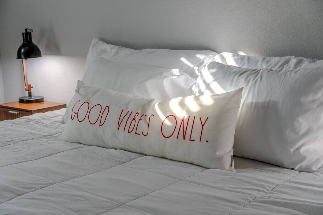 Una cama con ropa de cama blanca y un almohadón con la inscripción 'GOOD VIBES ONLY'.