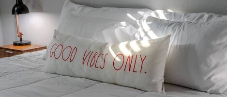 Una cama con ropa de cama blanca y un almohadón con la inscripción 'GOOD VIBES ONLY'.