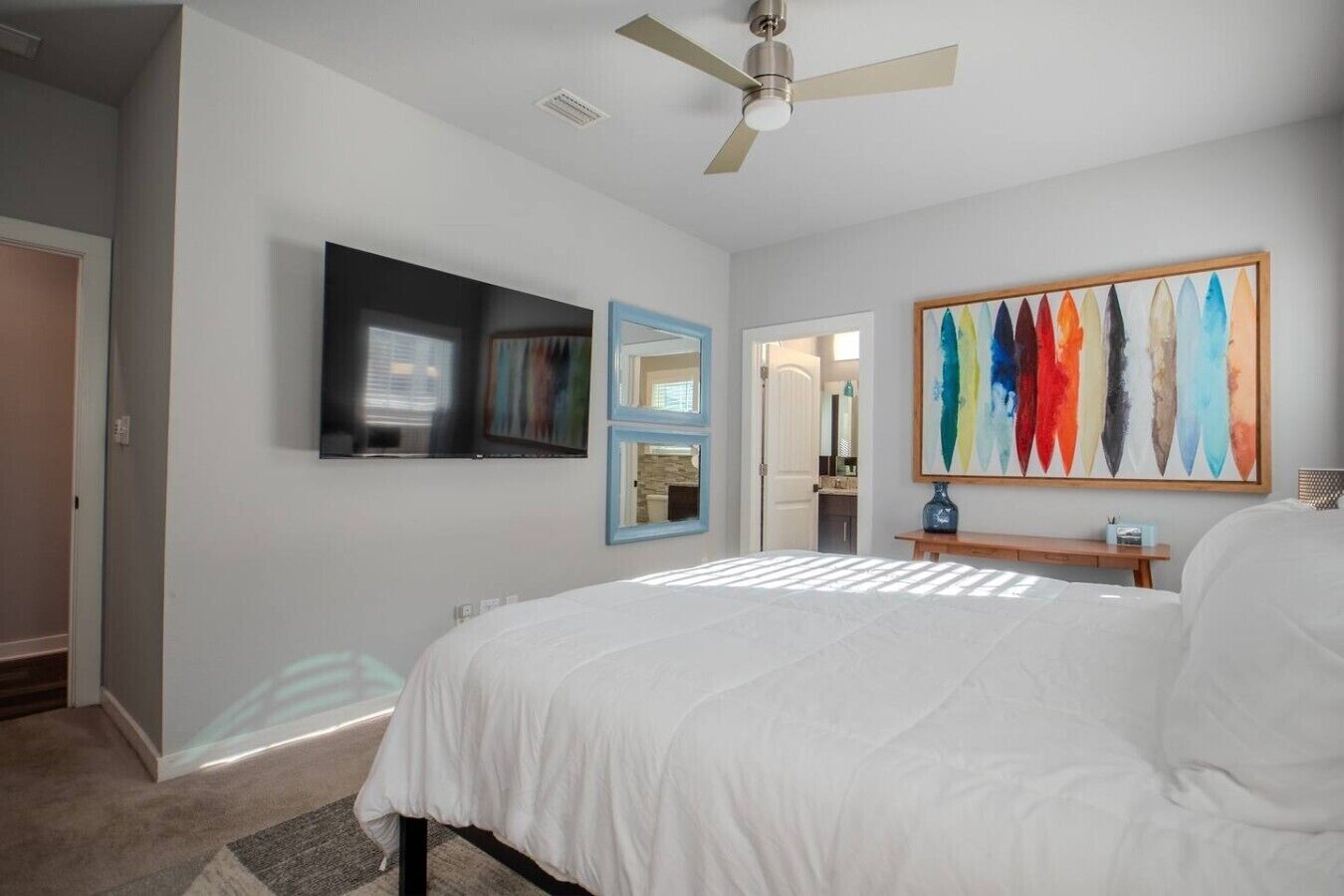 A bedroom with a bed, a ceiling fan, a TV, a painting, and a door leading to another room.