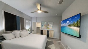 A hotel room with a bed, a ceiling fan, a large window, a painting of a beach, and a small kitchenette. - The Jazzy Junction-Great Downtown Location! (Pensacola)