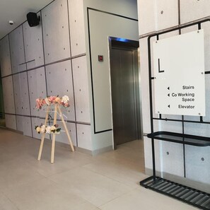 Un interior moderno con un arreglo floral sobre un pedestal, un ascensor y señalización direccional para las escaleras, un espacio de coworking y el ascensor.