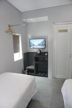 Habitación de hotel con dos camas, un televisor montado en la pared, perchero, pequeña cocina y una puerta.