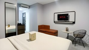 Una habitación de hotel moderna con una cama, un sillón de cuero marrón, una pequeña mesa redonda, una silla negra, un televisor montado en la pared que muestra 'NETFLIX' y un espejo.