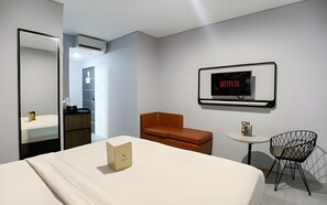 A modern hotel room with a bed, a brown leather chair, a small round table, a black chair, a wall-mounted TV displaying 'NETFLIX,' and a mirror. - Sambina Prime Hotel by Danapati (Jakarta)