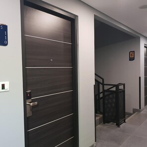 A modern hallway with a dark wooden door, a blue room number sign, and a staircase with a black metal railing. - Sambina Prime Hotel by Danapati (Jakarta)