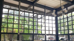 Un espacio al aire libre techado con sillas y una mesa, ventanas amplias con vistas a la vegetación y un ventilador de techo.