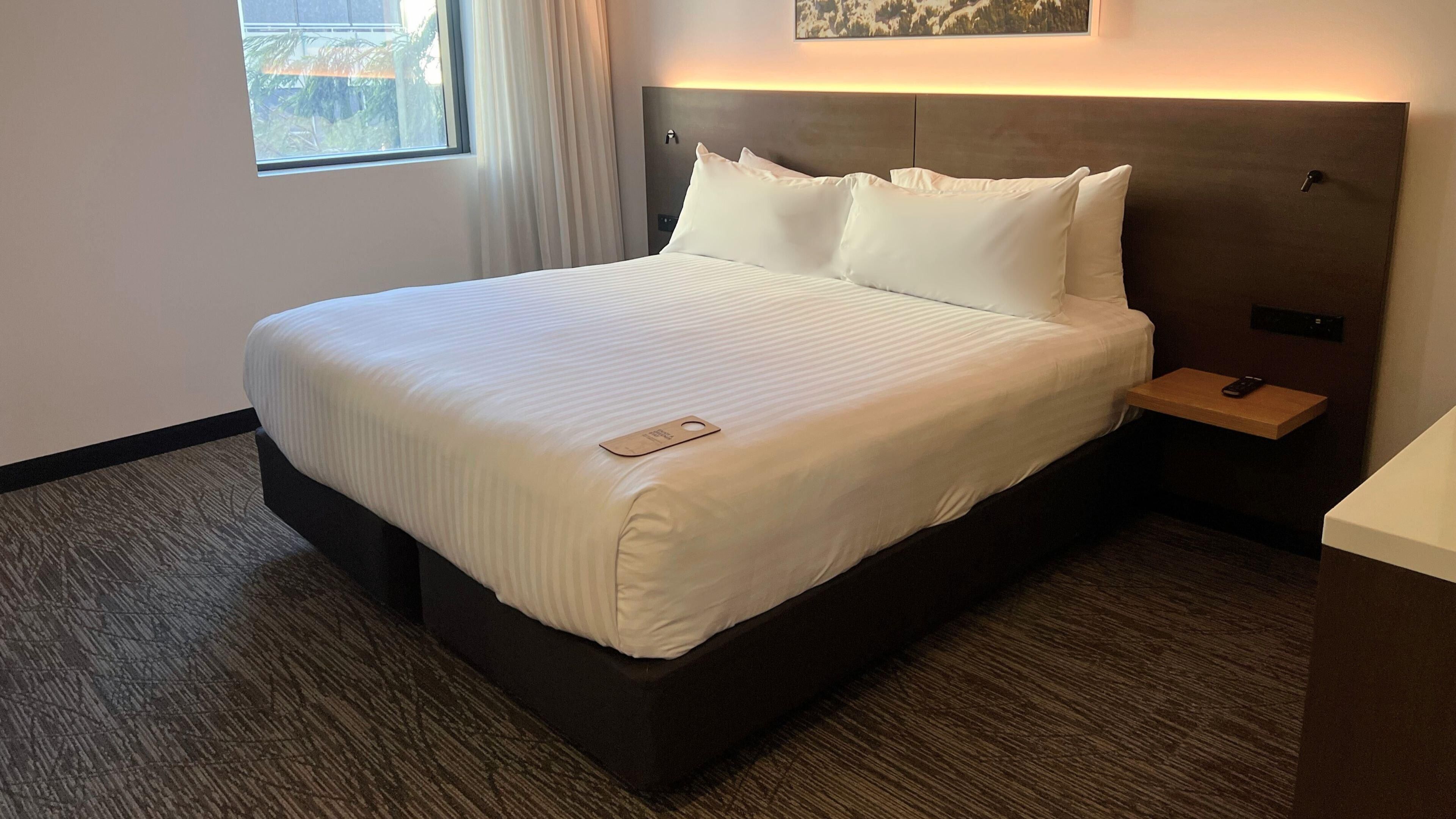 A hotel room with a large bed, a bedside table with a remote control, a nightstand, and a window with curtains.