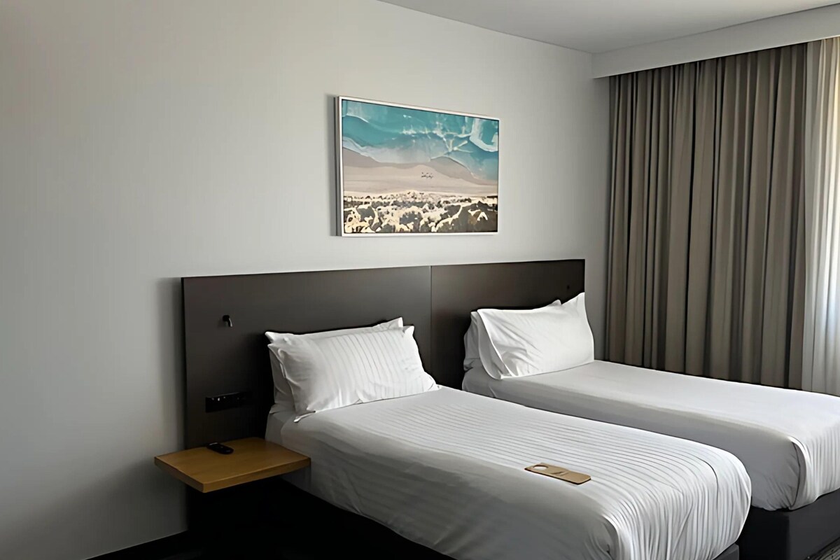 Habitación de hotel con dos camas, una mesita de noche, un control remoto, un cuadro de un paisaje desértico y un ventanal con cortinas.