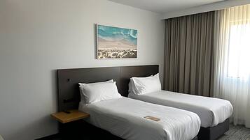 A hotel room with two beds, a nightstand, a remote control, a painting of a desert landscape, and a large window with curtains.