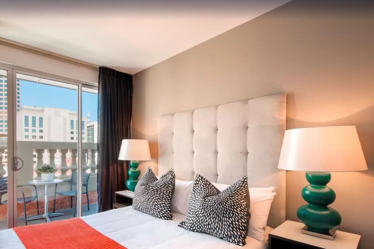 Habitación de hotel con una cama grande, dos lámparas de noche, un balcón con vistas a edificios y una manta roja.