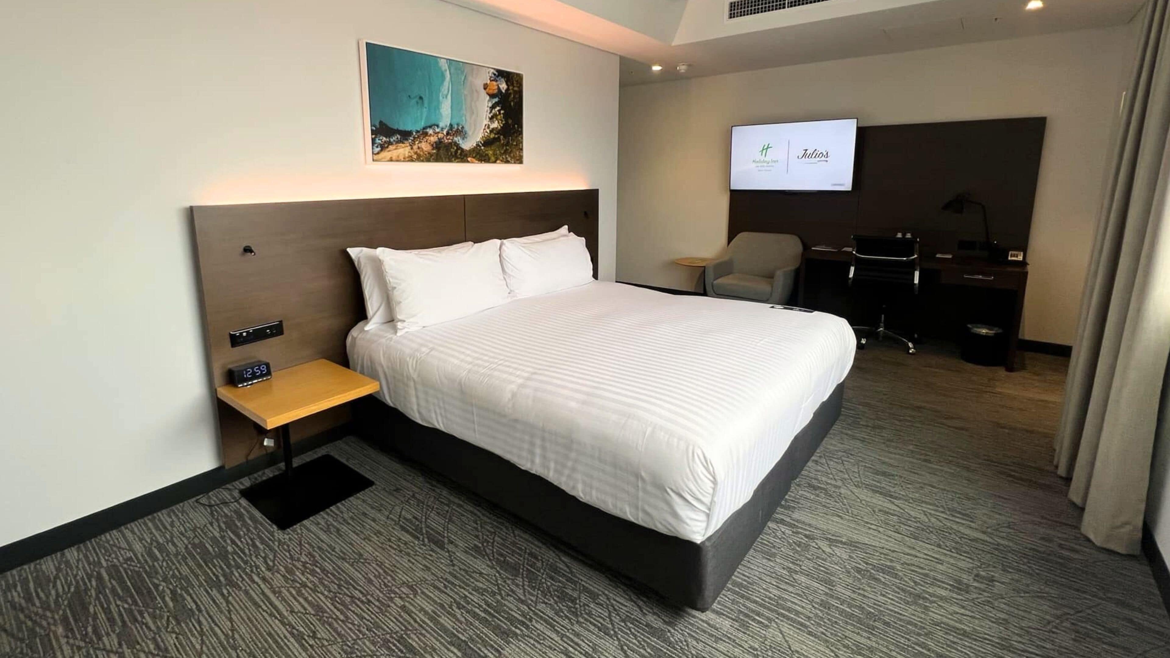 A hotel room with a large bed, a bedside table with a clock, a desk with a chair, and a TV screen displaying a hotel name.