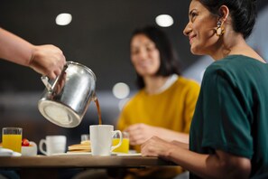 Eine Person gießt ein Getränk in eine Tasse an einem Tisch, an dem eine weitere Person sitzt.