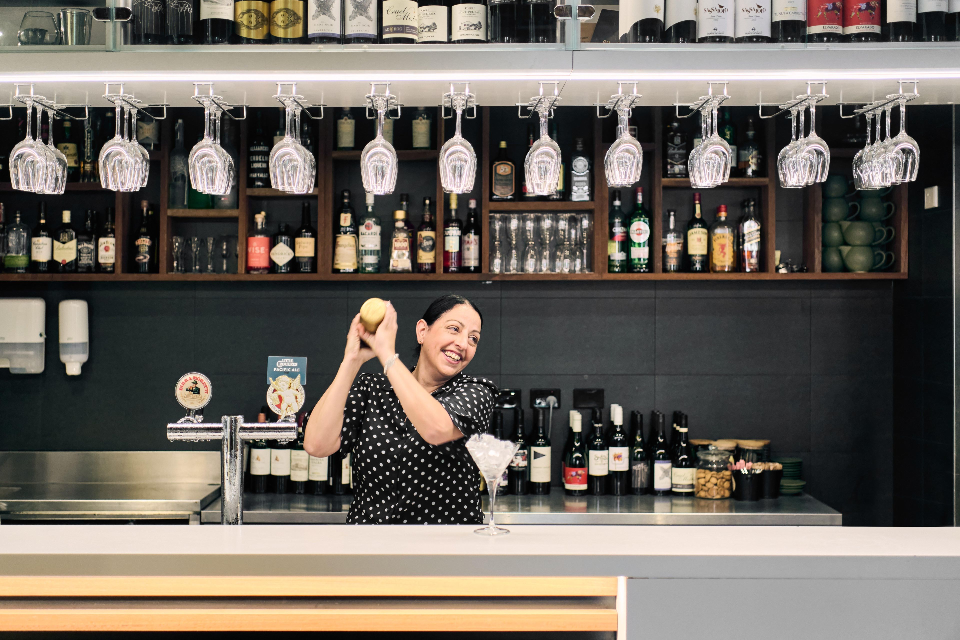 A bar area with shelves of liquor bottles, a bartender preparing a drink, and hanging glassware.