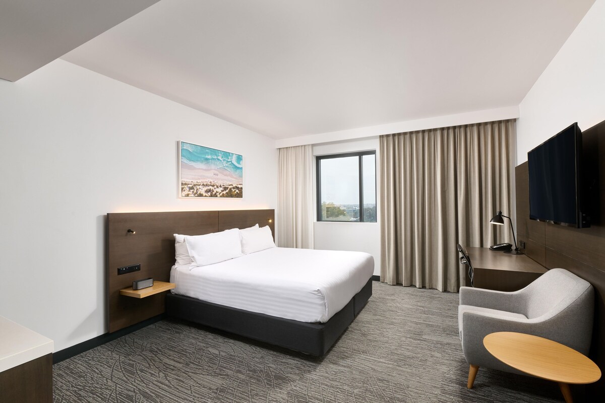 Habitación de hotel con cama, escritorio, silla, televisor y ventana con cortinas.
