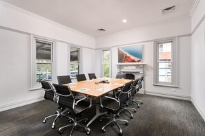 Uma sala de conferências com uma mesa grande, cadeiras pretas, paredes brancas e janelas amplas.