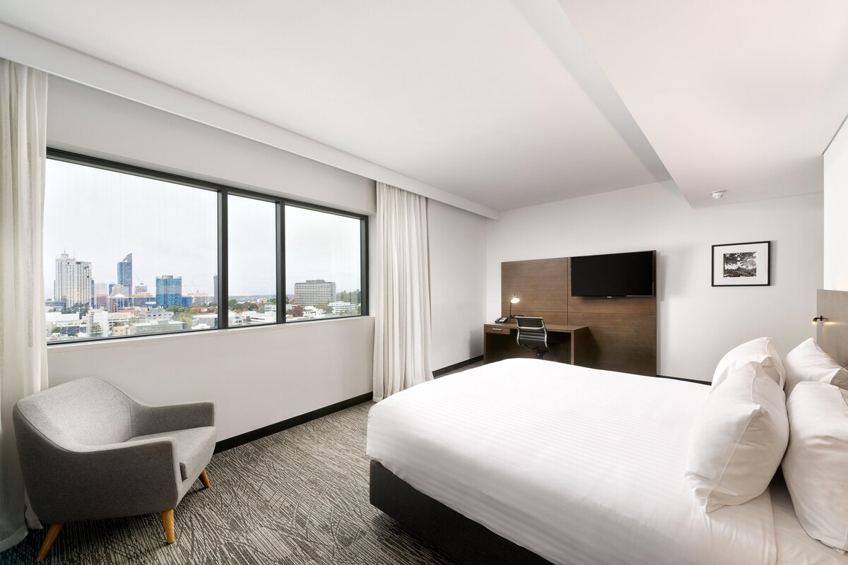 Habitación de hotel con un ventanal grande, cama, sillón, escritorio y televisor de pantalla plana.