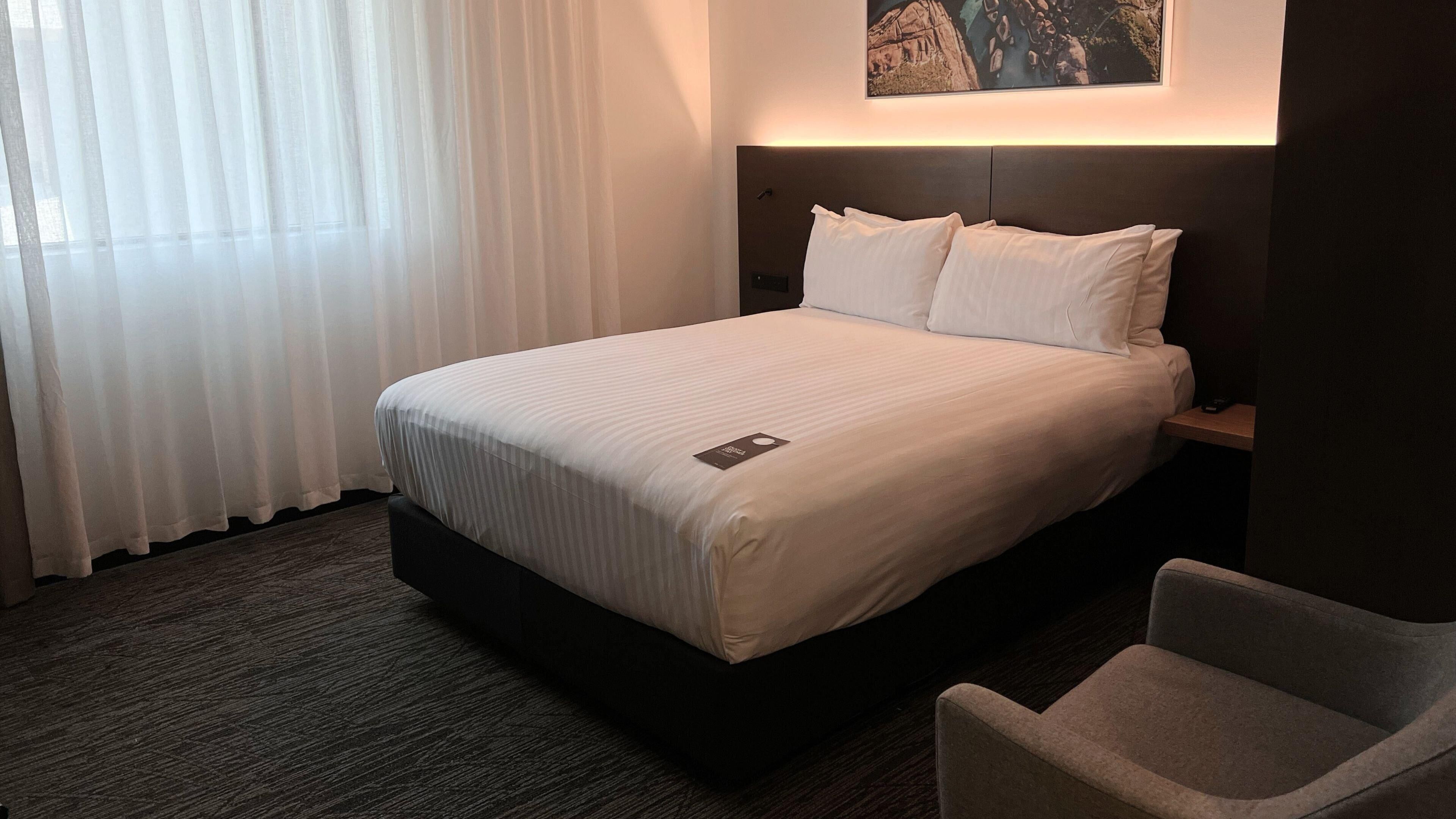 A hotel room with a bed, a nightstand, a chair, a painting on the wall, and a window with curtains.