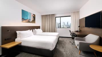 A hotel room with a bed, two bedside tables, a desk, a chair, a TV, and a large window with a city view.