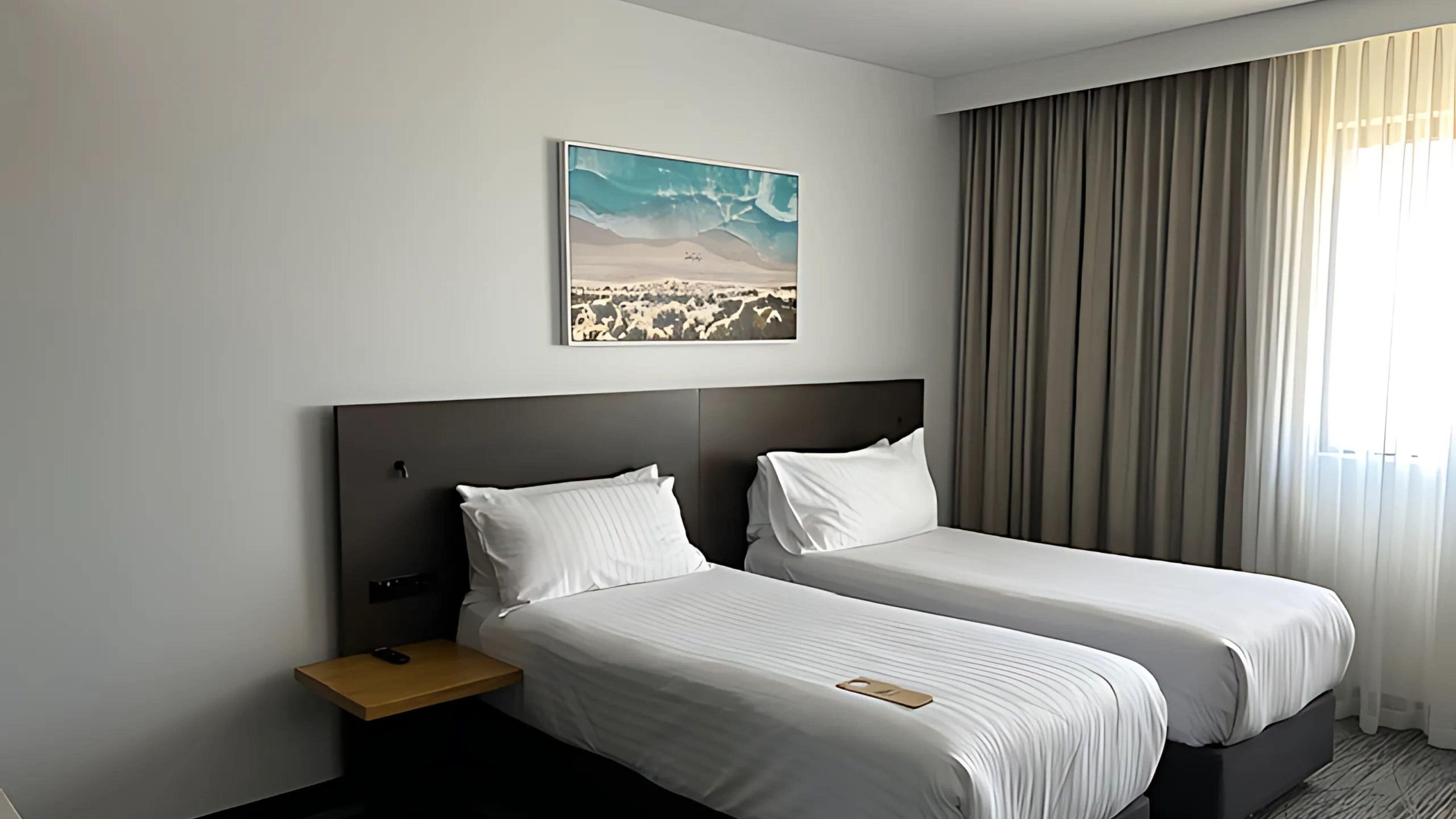A hotel room with two beds, a nightstand, a remote control, a painting of a desert landscape, and a large window with curtains.
