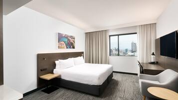 A hotel room with a bed, a desk, a chair, a TV, a window with curtains, and a city view.