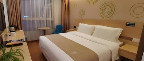 A hotel room with a bed, bedside table, chair, desk, and a potted plant.