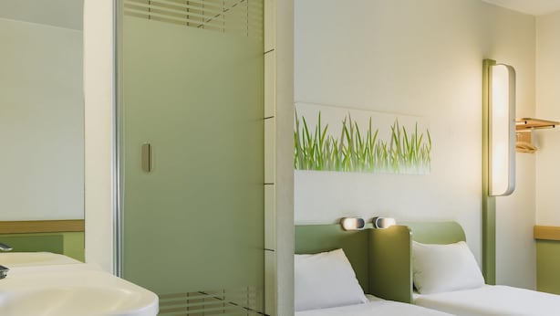 Habitación de hotel con cabina de ducha de vidrio, lavamanos con toalla, dos camas con ropa blanca y un aplique de pared.