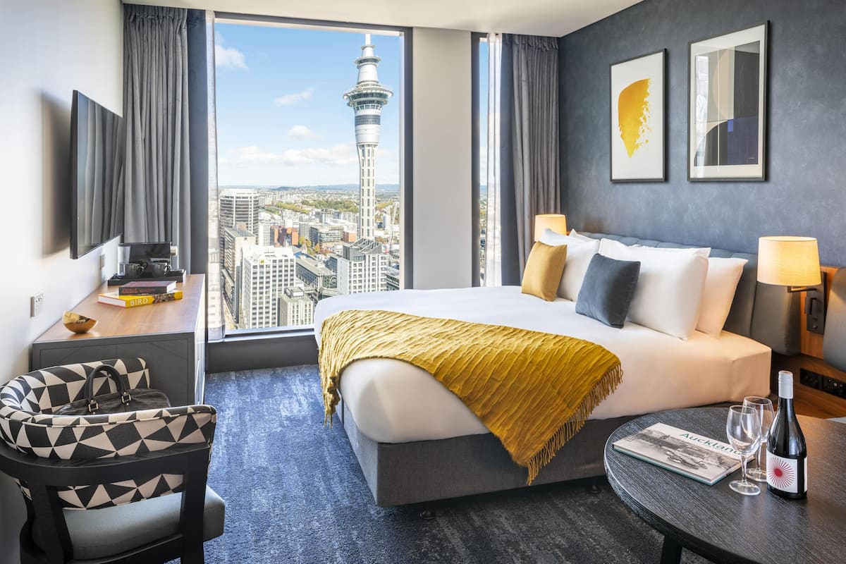 A hotel room with a bed, a chair, a table with a bottle and glasses, a TV, and a view of a cityscape with a tower.
