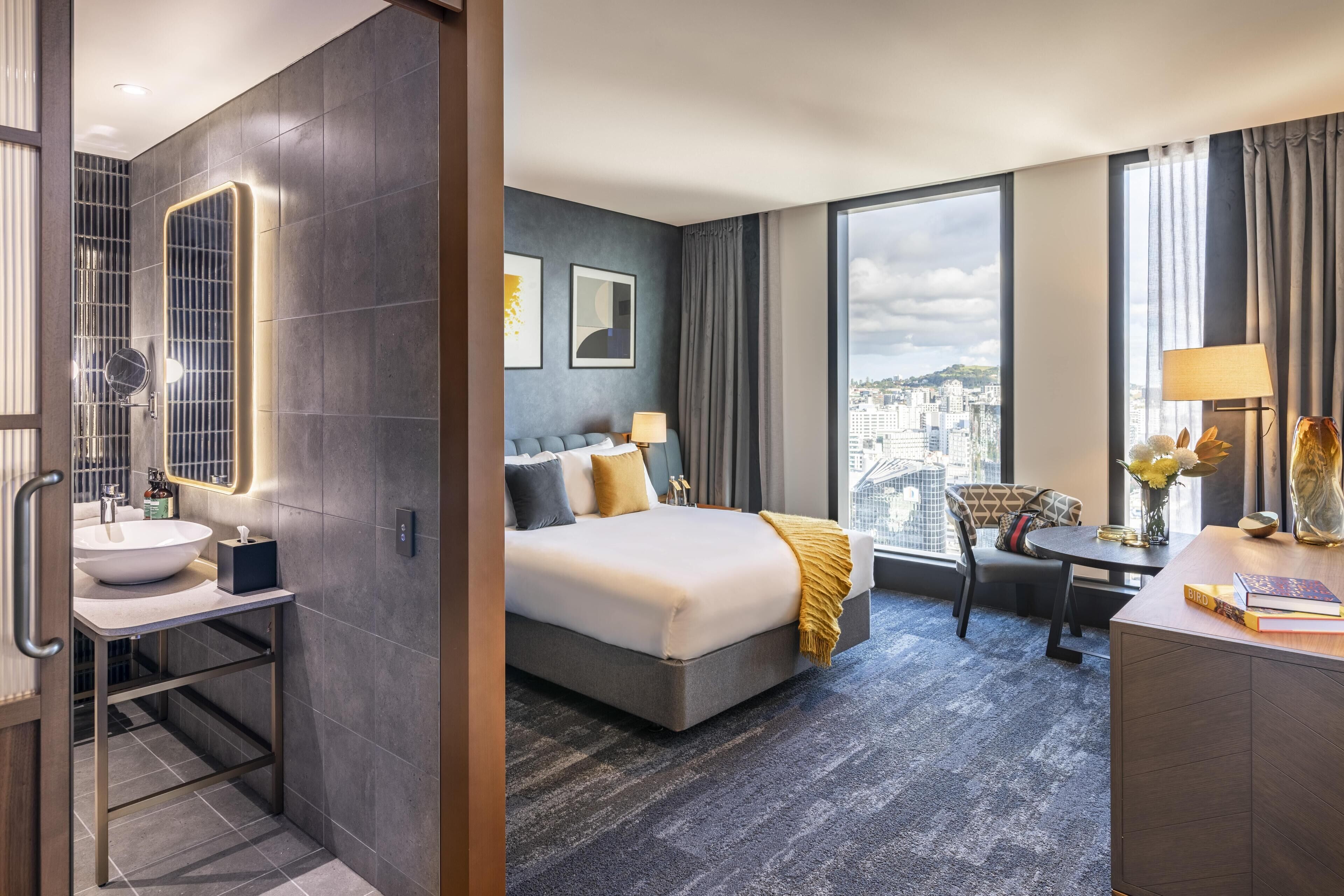A hotel room with a large bed, a desk, a chair, a bathroom with a sink, and a large window with a city view.