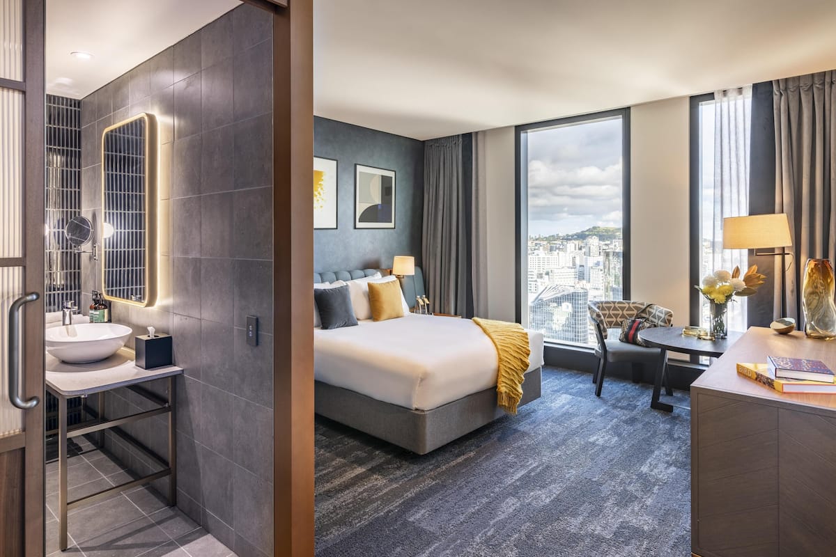 A hotel room with a large bed, a desk, a chair, a bathroom with a sink, and a large window with a city view.