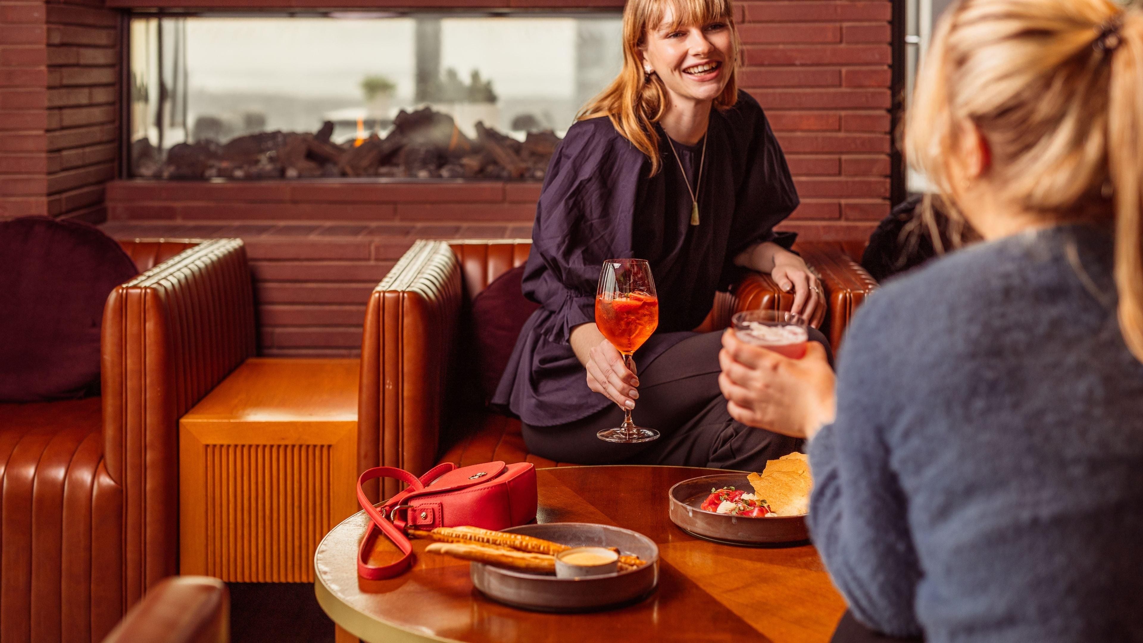 A person sitting on a leather couch with a glass of drink, a red bag, and a plate of food on a table.
