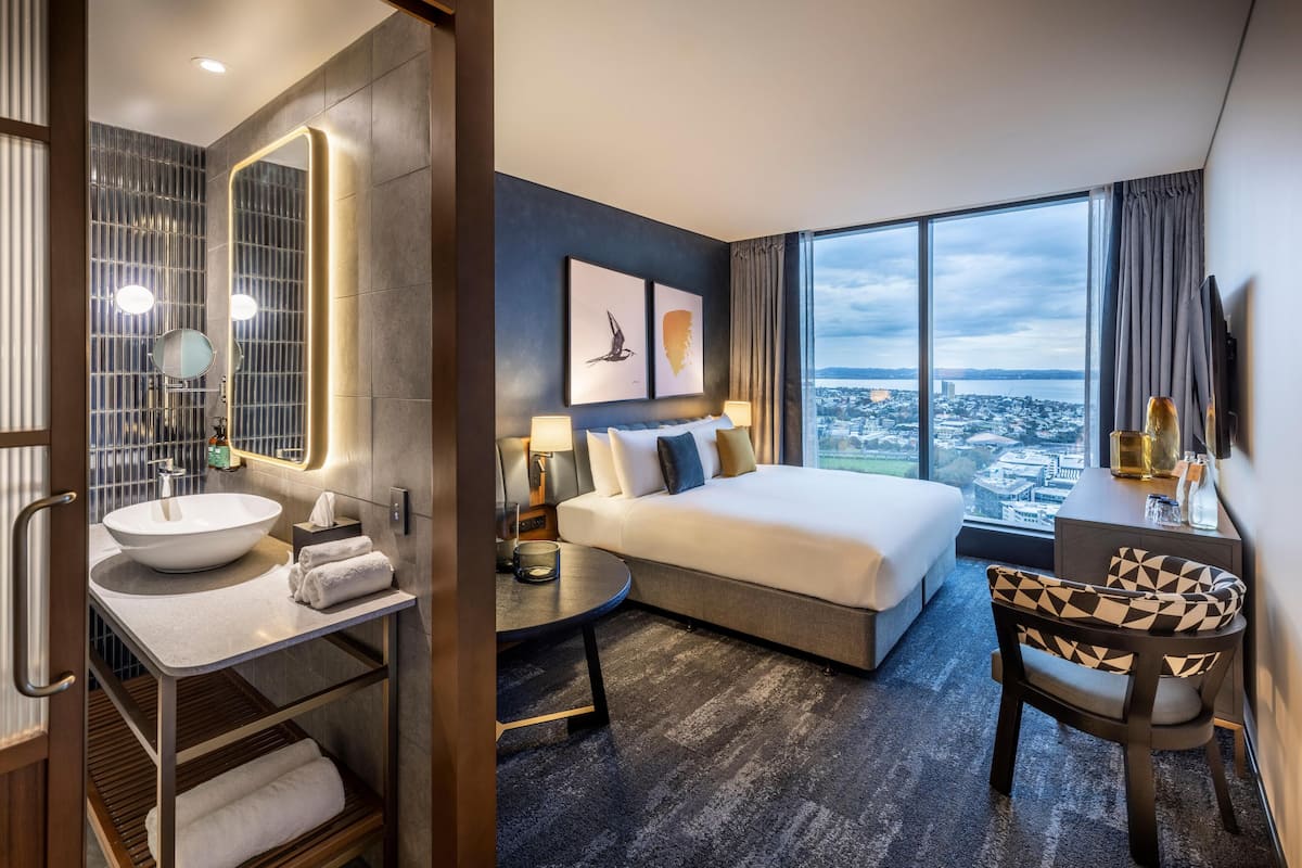 A hotel room with a large bed, a bathroom with a sink and mirror, a small table, a chair, and a city view.