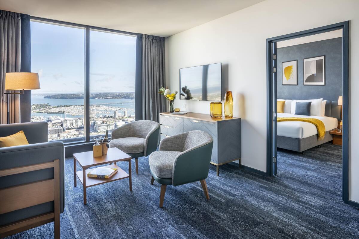 A hotel room with a large window, a desk, two chairs, a bed, and a mirror.