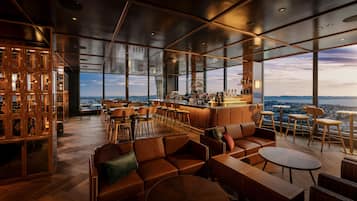 Uma área de bar moderna, com pé direito alto, grandes janelas e vista para a paisagem urbana.