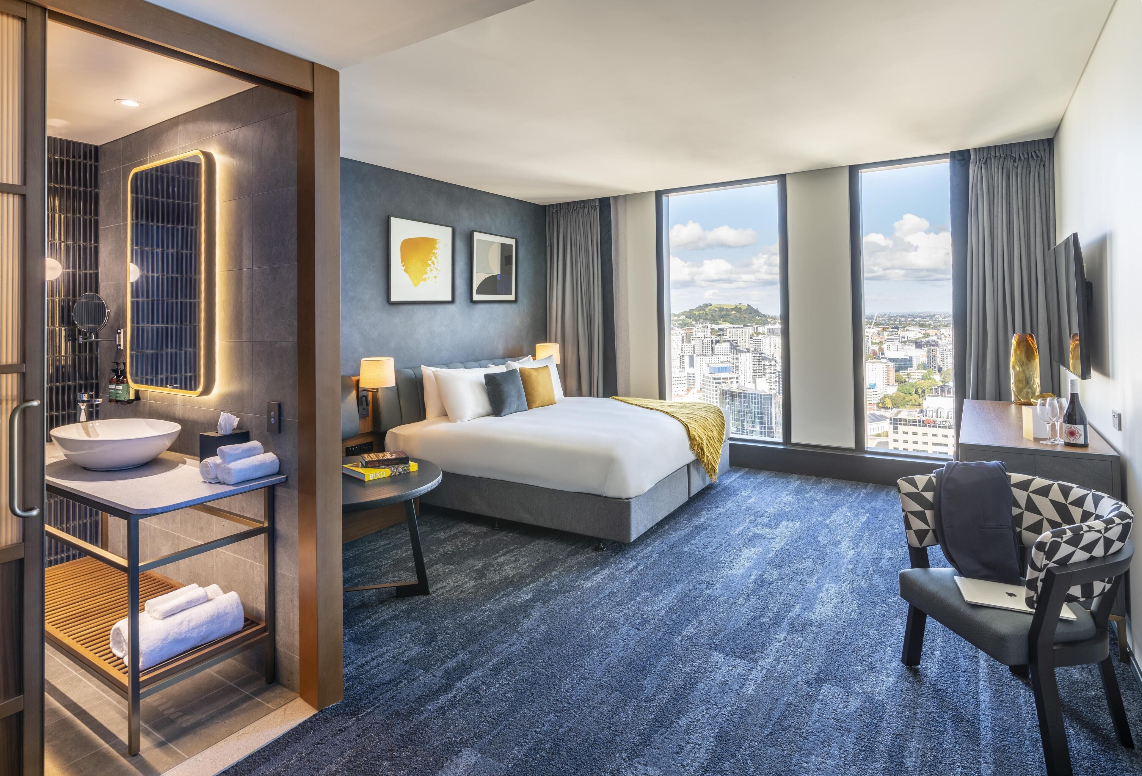A modern hotel room with a large bed, a desk, a chair, a bathroom with a sink and mirror, and a city view.