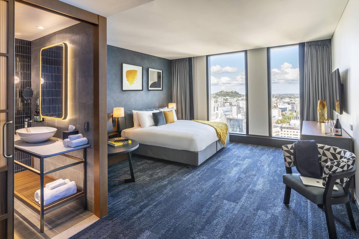 A modern hotel room with a large bed, a desk, a chair, a bathroom with a sink and mirror, and a city view.