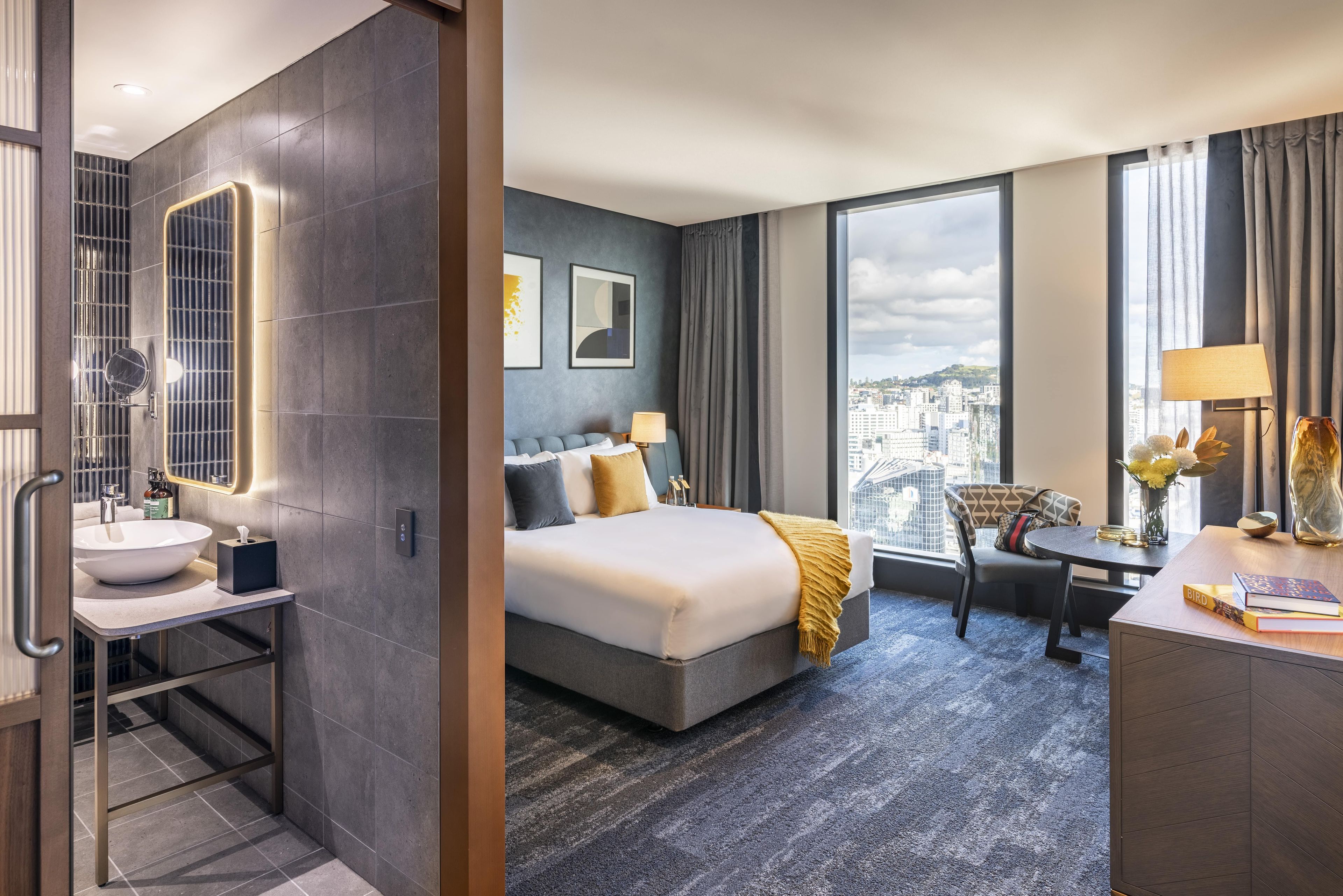 Une chambre d’hôtel avec un grand lit, un bureau, une chaise, une salle de bain avec un lavabo et une grande fenêtre donnant sur la ville.
