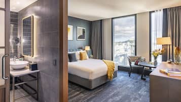 Habitación de hotel con una cama grande, un escritorio, una silla, un baño con lavamanos y un ventanal con vistas a la ciudad.