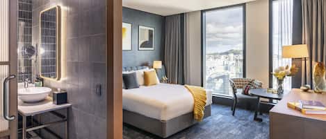A hotel room with a large bed, a desk, a chair, a bathroom with a sink, and a large window with a city view.