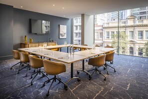 A conference room with a long table, chairs, a flat-screen TV, artwork, and a view of buildings through large windows. - Voco Auckland City Centre (Auckland)